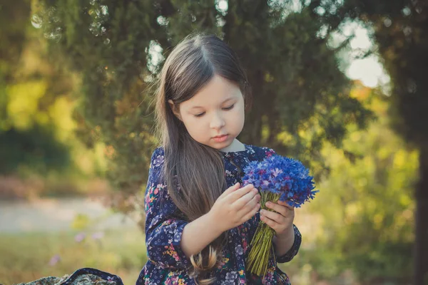 Bébé fille mignonne avec des cheveux bruns et des yeux marron portrait avec des fleurs sauvages pourpres bleu profond pieds nus portant robe de fleur colorée élégante et regardant à la caméra avec le visage brillant — Photo