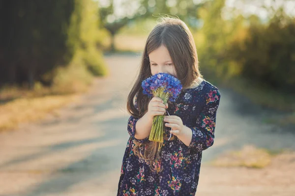 Menina bonito com cabelos morena e olhos castanhos retrato com azul profundo roxo flores selvagens descalço vestindo elegante vestido de flor colorido e olhando para a câmera com rosto brilhante — Fotografia de Stock