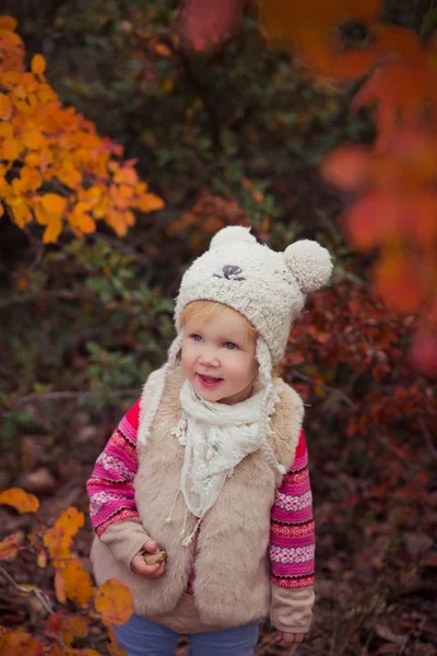 Schattige jonge Russische babymeisje stijlvol gekleed in warm wit bont handgemaakte jas blauw jeans laarzen en hoed teddybeer poseren in de herfst kleurrijk bos traject gezicht met roze Wang verslaafd — Stockfoto