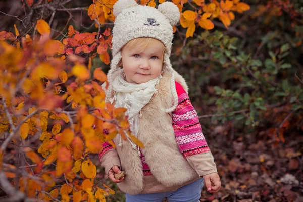 Милий молодий російський дівчинку стильний одягнені в теплий білий жакет ручної роботи сині джинси Унти і підключили капелюх плюшевого ведмедика постановки в осінній барвистий ліс шлях обличчя з рожеві щоки — стокове фото