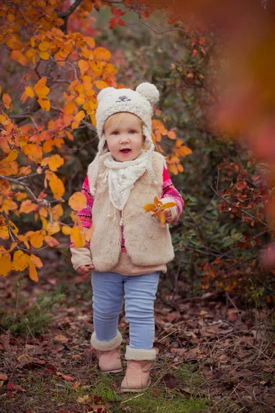 Schattige jonge Russische babymeisje stijlvol gekleed in warm wit bont handgemaakte jas blauw jeans laarzen en hoed teddybeer poseren in de herfst kleurrijk bos traject gezicht met roze Wang verslaafd — Stockfoto