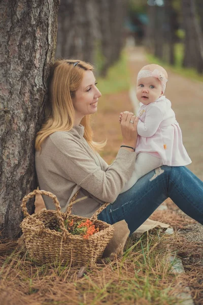 快乐的年轻母亲 sylish 打扮与女婴穿着粉红色的礼服可爱的花一生一起坐在森林附近的树木和篮子与野生浆果 — 图库照片