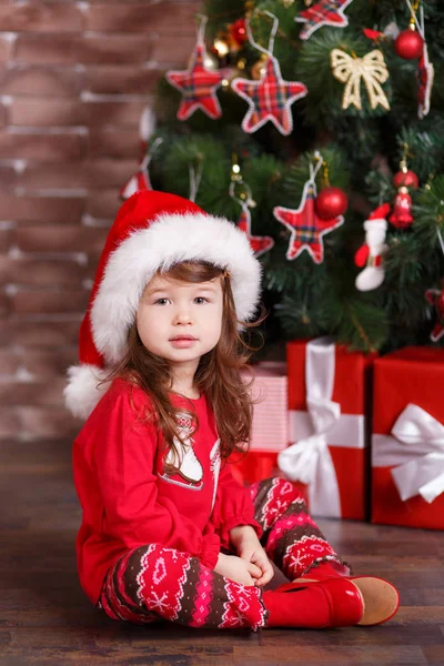 Jovem morena boneca menina elegante vestida de camisola pulôver vermelho quente sox sapatos chapéu de Santa sorrindo posando sentado em estúdio inverno árvore de natal com presentes com lábios de faneca e bochechas rosa . — Fotografia de Stock