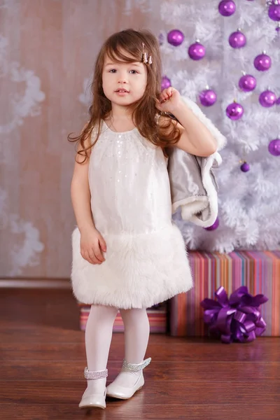Молода брюнетка лялька леді стильно одягнена в затишному вечірньому офіційному білому платті куртка ремінець взуття посміхається позує в студії зимової різдвяної ялинки і подарунки з пухкими губами і рожевими щоками . — стокове фото
