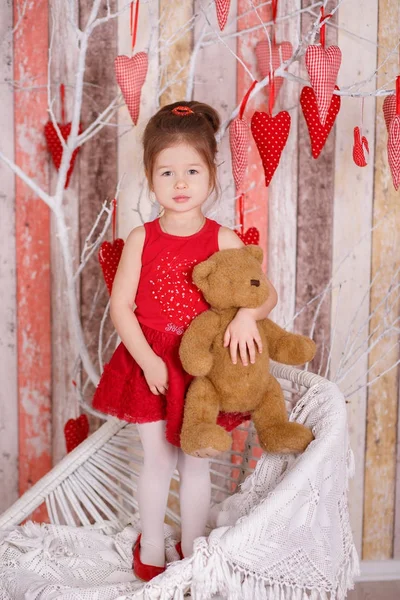 Young brunette dolly lady girl stylish dressed in red dress strap shoes smiling posing sitting in studio on white chair with teddy bear and tree with hearts with pout lips and pink cheeks. — Stock Photo, Image