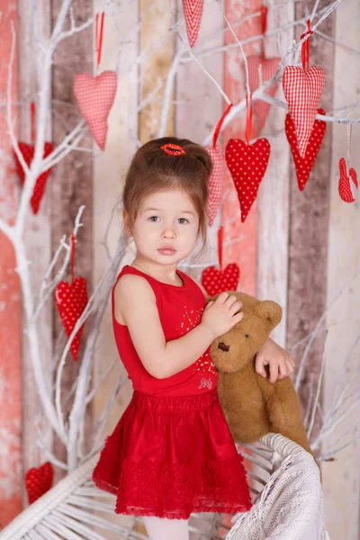 Jovem morena dolly senhora menina elegante vestida com sapatos de alça de vestido vermelho sorrindo posando sentado em estúdio na cadeira branca com ursinho de pelúcia e árvore com corações com lábios de faneca e bochechas rosa . — Fotografia de Stock