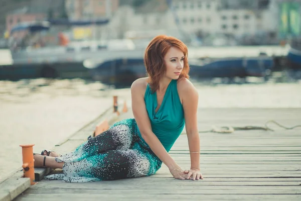 Increíble hermosa dama chica mujer con el pelo rojo llama usando ropa verde de moda de lujo posando sentado en embarcadero de madera embarcadero wth puerto barcos de fondo Adorable escena de sirena de mar — Foto de Stock