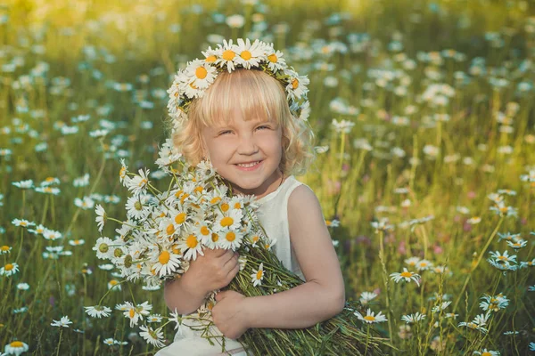 Mignon jeune fille blonde en robe blanche aérée posant sur la marguerite prairie de camomille avec bouquet de fleurs dans le panier des mains seau en métal et couronne anademon tête supérieure souriante . — Photo