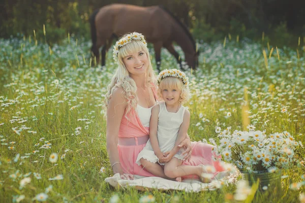 Magnifique image de blonde adorable dame mère mère mère en robe rose aérée avec fille fille bébé en blanc ambrace l'un l'autre sur marguerite prairie de camomille avec cheval sur fond . — Photo