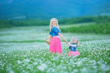 Anne ve kızı bir piknik papatya alanındaki tarih. Kot ceket ve pembe giyen iki güzel sarışın anne etek ve küçük kız papatya alanında bulunan buket dandalion sepet kucaklayan