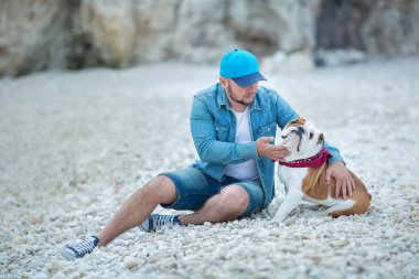 Yakışıklı kot adam güzel İngilizce bulldog ile birlikte bir plaj deniz tarafında oturan Mavi şapkalı kalın şık giyinmiş.