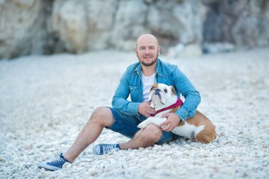Yakışıklı kot adam güzel İngilizce bulldog ile birlikte bir plaj deniz tarafında oturan Mavi şapkalı kalın şık giyinmiş.