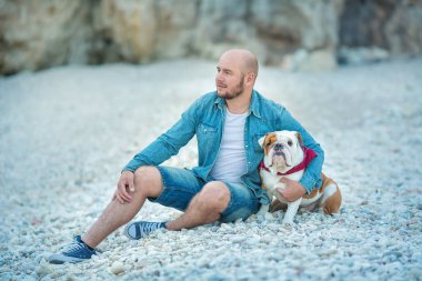 Yakışıklı kot adam güzel İngilizce bulldog ile birlikte bir plaj deniz tarafında oturan Mavi şapkalı kalın şık giyinmiş.