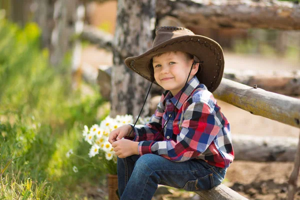 Niedlichen Fermer gutaussehenden Jungen Cowboy in Jeans genießen Sommertag im Dorfleben mit Blumen tragen ledernen Kuhhut glücklich lächelnd. — Stockfoto