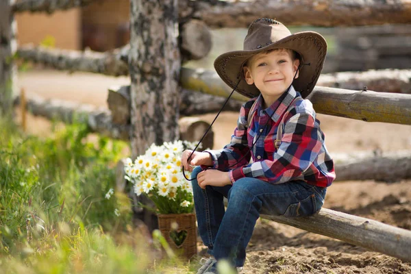 Niedlichen Fermer gutaussehenden Jungen Cowboy in Jeans genießen Sommertag im Dorfleben mit Blumen tragen ledernen Kuhhut glücklich lächelnd. — Stockfoto