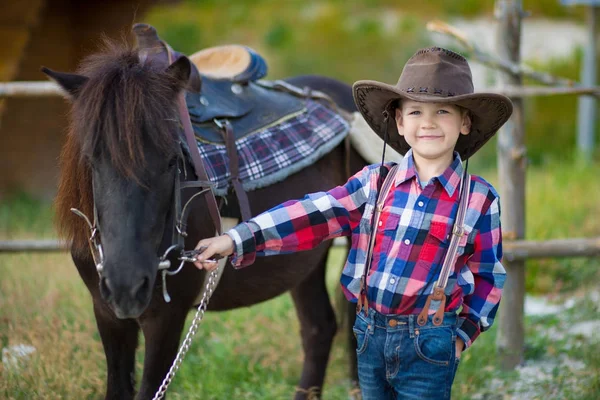 Schattig fermer knappe jongen cowboy in spijkerbroek genieten van zomerdag in het dorpsleven met bloemen dragen lederen koe hoed happyly glimlachend en zittend op paard . — Stockfoto