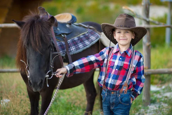 Cute fermer handsome boy cowboy in jeans enjoying summer day in village life with flowers wearing leather cow hat happyly smiling and sitting on horse . — Stock Photo, Image