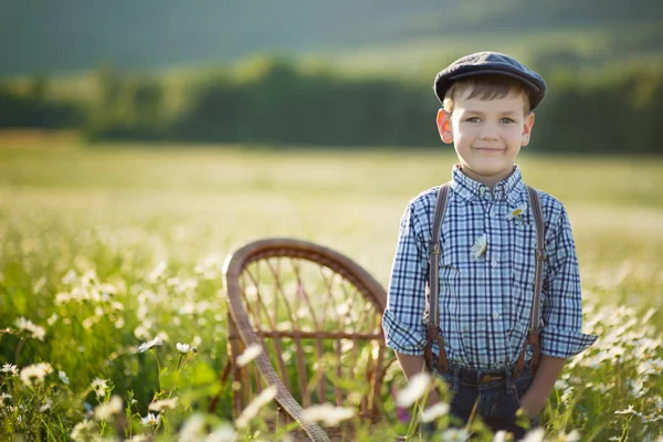 Netter gutaussehender Junge in Jeans und Hemd sitzt auf einem Holzstuhl auf einer Kamillenwiese und genießt lächelnd den Sommertag auf einem Bauernhof. — Stockfoto