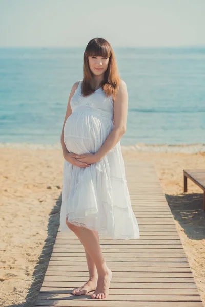 Справжня мила вагітна леді-жінка в білому повітряному платті, що йде піщаним пляжем, дерев'яна палітра, що тримає животик. Приваблива красива молода дівчина насолоджується теплим літом з ненародженою дитиною в животі . — стокове фото