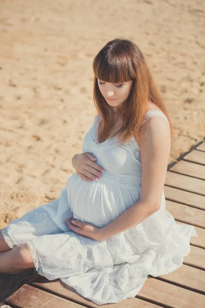 Справжня мила вагітна леді-жінка в білій повітряній сукні, що сидить на піщаному пляжі дерев'яний палітральний міст, що тримає животик. Приваблива красива молода дівчина насолоджується теплим літом з ненародженою дитиною в животі . — стокове фото