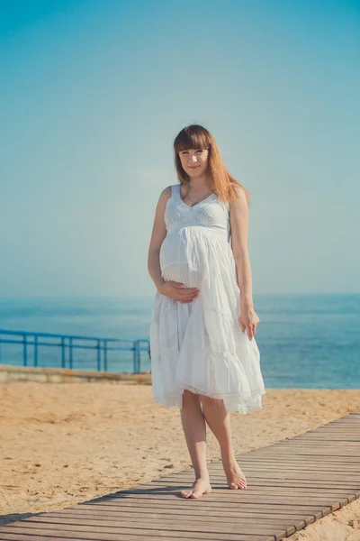 Справжня мила вагітна леді-жінка в білому повітряному платті, що йде піщаним пляжем, дерев'яна палітра, що тримає животик. Приваблива красива молода дівчина насолоджується теплим літом з ненародженою дитиною в животі . — стокове фото