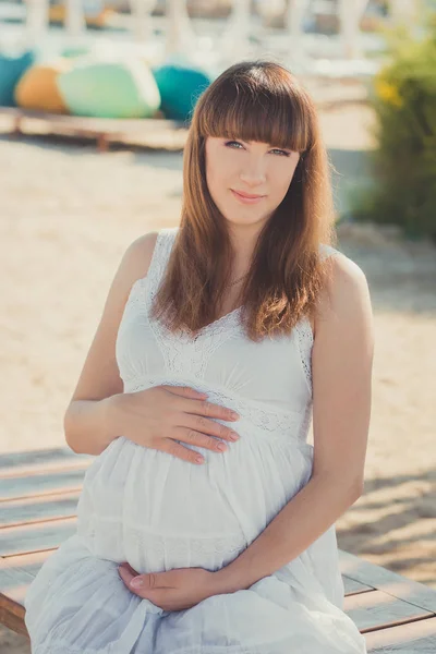 Справжня мила вагітна леді-жінка в білій повітряній сукні, що сидить на піщаному пляжі дерев'яний палітральний міст, що тримає животик. Приваблива красива молода дівчина насолоджується теплим літом з ненародженою дитиною в животі . — стокове фото