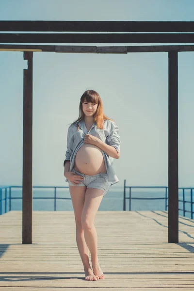 Genuine cute pregnant lady woman in jeans shirt posing beach wooden palette bridge holding tummy abdomen dreaming. Attractive beautiful young girl enjoying warm summer with unborn child in belly. — Stock Photo, Image