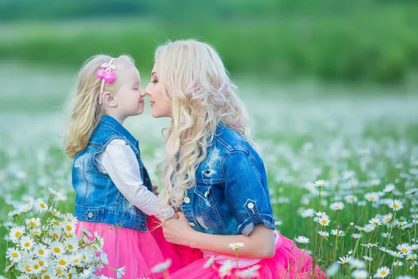 Mom and daughter on a picnic in the chamomile field. Two beautiful blondes wearing jeans jacket and pink skirt Mother and baby girl embracing in the chamomile field with basket of bouquet dandalion — Stock Photo, Image