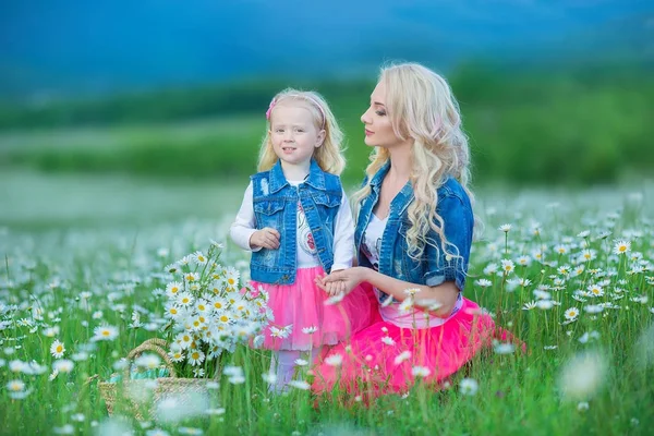 Máma a dcera na piknik v poli heřmánek. Dvě krásné blondýnky na sobě džínové bundy a růžová sukně matka a dítě dívka objímá v poli heřmánek s košíkem kytice dandalion — Stock fotografie