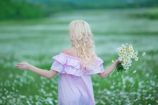 Atractiva rubia en el campo de manzanilla. Mujer joven en corona rodeada de manzanillas con elegante vestido rosa disfrutando de la primavera de verano —  Fotos de Stock