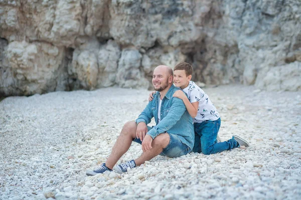 Guapo audaz elegante vestido en jeans hombre en gorra azul sentado en un lado del mar de playa junto con lindo hijo adolescente andpretty bulldog inglés . — Foto de Stock