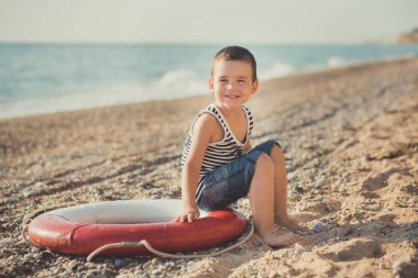 Çizgili bir gömlek ile deniz lifebuoy liferound plajında bir çocuk oturur. Bir çocuk kum güzel bir plaj üzerine oturur. Güzel bir plaj ve deniz