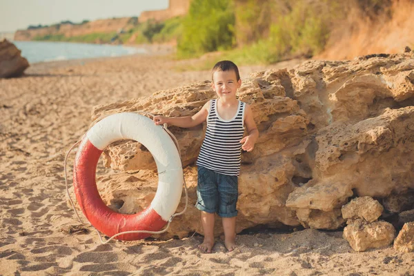 En pojke sitter på stranden i en randig tröja med en marin livboj, liferound. En pojke sitter på sanden på en vacker strand. Vacker strand och havet — Stockfoto