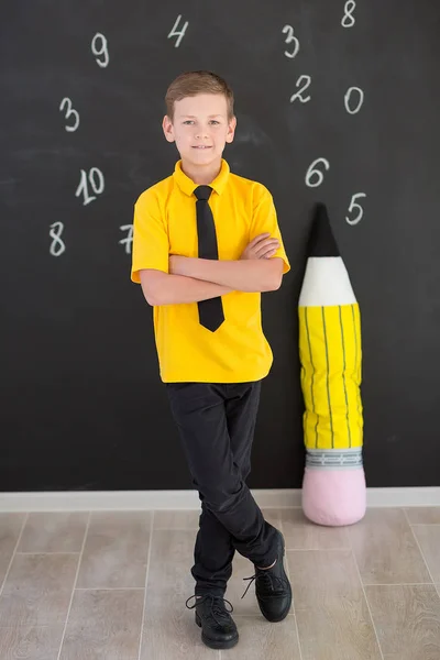 Cute handsome school boy in yellow t-shirt tie and stylish boots casuals standing cloase to black board with numbers and holding huge big soft toy pencil and smiling