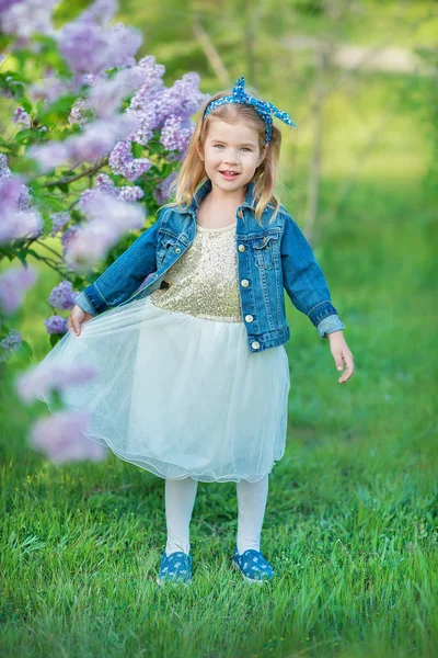 可爱的女孩在蓝色夹克与仙女透气的裙子站立接近的丁香灌木 — 图库照片