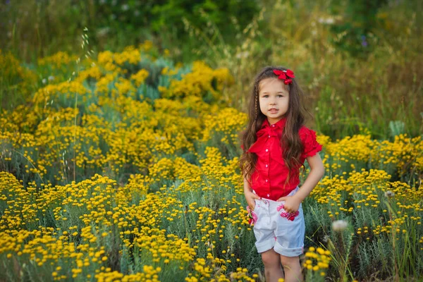 Jeune fille mignonne en chemise rouge et short blanc debout près de fleurs jaunes . — Photo
