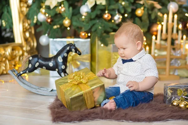 Beautiful little baby boy celebrates Christmas. New Year's holidays. Baby in a Christmas costume casual clothes with gifts on fur close to new year tree in studio decorations.