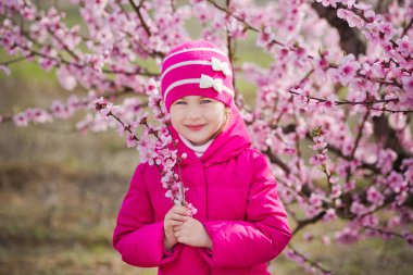 Güzel şık giyimli esmer ile şirin kız pembe çiçekli bahar genç şeftali ağacına sahasında duran anne anne. Bayan yağmur giyim ve mavi şapka lastik çizme giymiş bahar mevsimi.
