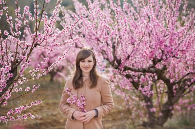 Güzel şık giyimli esmer ile şirin kız pembe çiçekli bahar genç şeftali ağacına sahasında duran anne anne. Bayan yağmur giyim ve mavi şapka lastik çizme giymiş bahar mevsimi.