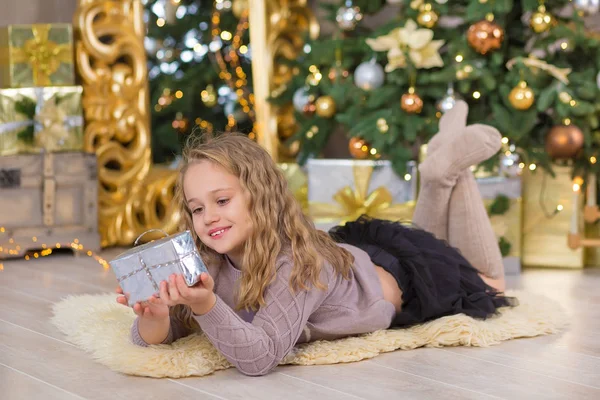 Mooi meisje glimlachend, zittend in de buurt van enorme gouden spiegel genoeg presenteert op bont kerst groen wit luxe boom decoraties en beneden liggen geschenken — Stockfoto