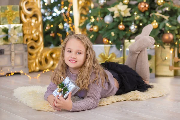 Mooi meisje glimlachend, zittend in de buurt van enorme gouden spiegel genoeg presenteert op bont kerst groen wit luxe boom decoraties en beneden liggen geschenken — Stockfoto