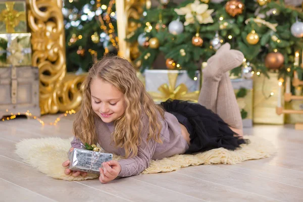 Mooi meisje glimlachend, zittend in de buurt van enorme gouden spiegel genoeg presenteert op bont kerst groen wit luxe boom decoraties en beneden liggen geschenken — Stockfoto