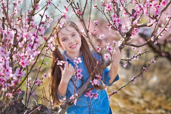 Bonito bonita elegante vestido morena e loiras meninas irmãs de pé em um campo de primavera jovem pêssego árvore com flores rosadas.Senhora vestida de jeans na temporada de primavera com sua irmã em vestido rosa . — Fotografia de Stock