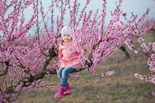 Cute beautiful stylish dressed brunette girl with mother mom standing on a field of spring young peach tree with pink flowers.Lady dressed in rain clothes and blue hat rubber boots spring season. — Stock Photo, Image