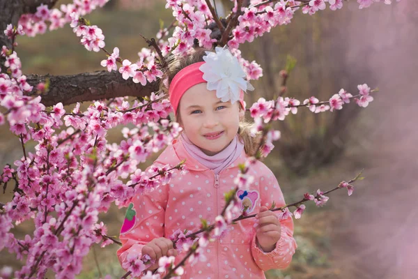 Cute beautiful stylish dressed brunette girl with mother mom standing on a field of spring young peach tree with pink flowers.Lady dressed in rain clothes and blue hat rubber boots spring season. Stock Photo