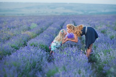 Lavanta sahada gülümseyen genç kızı ile genç anne. Kızı anne eller üzerinde oturuyor. Renkli elbise ve koyu mavi elbiseli anne kız.