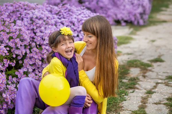 Gyönyörű szőke anya-anya és aranyos mosolygós lány lány élvezi időt együtt közel lila virágok színes ruhát visel. — Stock Fotó