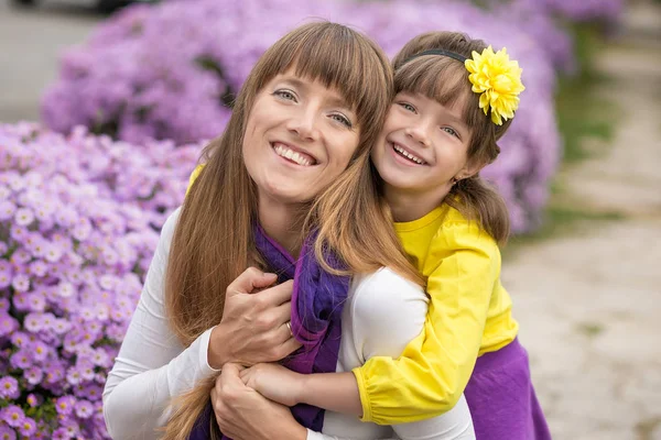 Gyönyörű szőke anya-anya és aranyos mosolygós lány lány élvezi időt együtt közel lila virágok színes ruhát visel. — Stock Fotó
