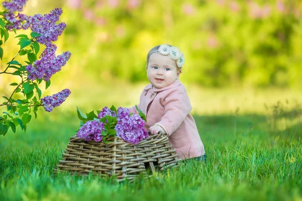 Smiling baby girl 1-2 year old wearing flower wreath, holding bouquet of lilac outdoors. Looking at camera. Summer spring time. — Stock Photo, Image