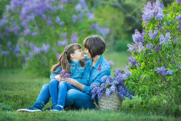 Bonito adorável linda mãe senhora mãe mulher com morena menina filha no prado de lilás roxo bush.People em jeans desgaste . — Fotografia de Stock